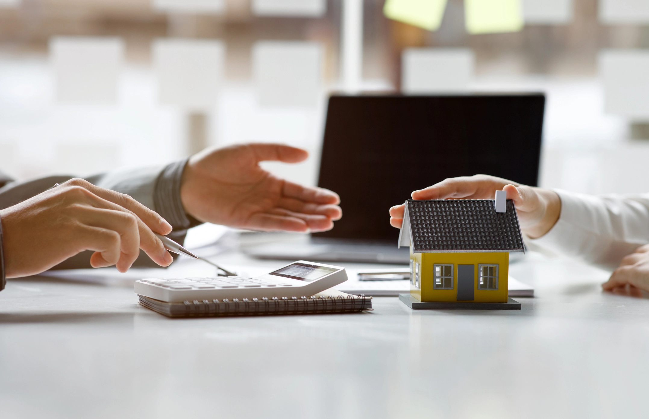 Hands discussing a home loan with a model house and calculator on a desk, symbolizing financial planning and support for homeownership